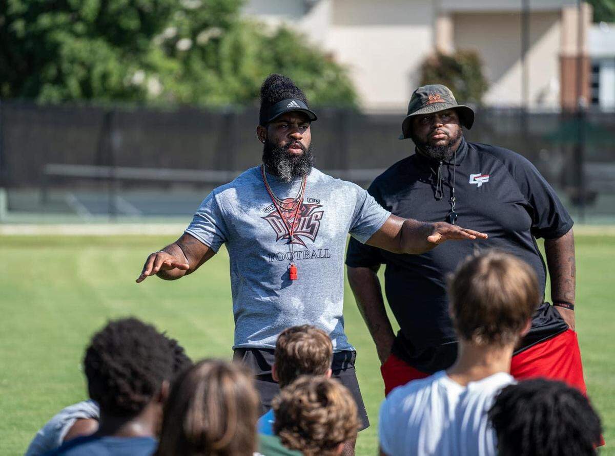 Great Falls head coach Syvelle Newton (middle, grey shirt) and defensive coordinator Tyler Hicks (right, black shirt) address their players at practice.