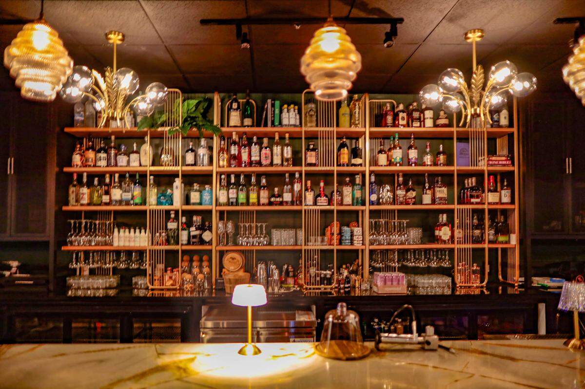 Looking across a stunning gold-and-green veined marble bar top, the view is of a large, well-stocked back bar. The wooden shelves are warmly lit, highlighting hundreds of liquor bottles and hanging glassware. The space is lit by several ornate, modern chandeliers and pendant lights, creating an upscale, intimate atmosphere.