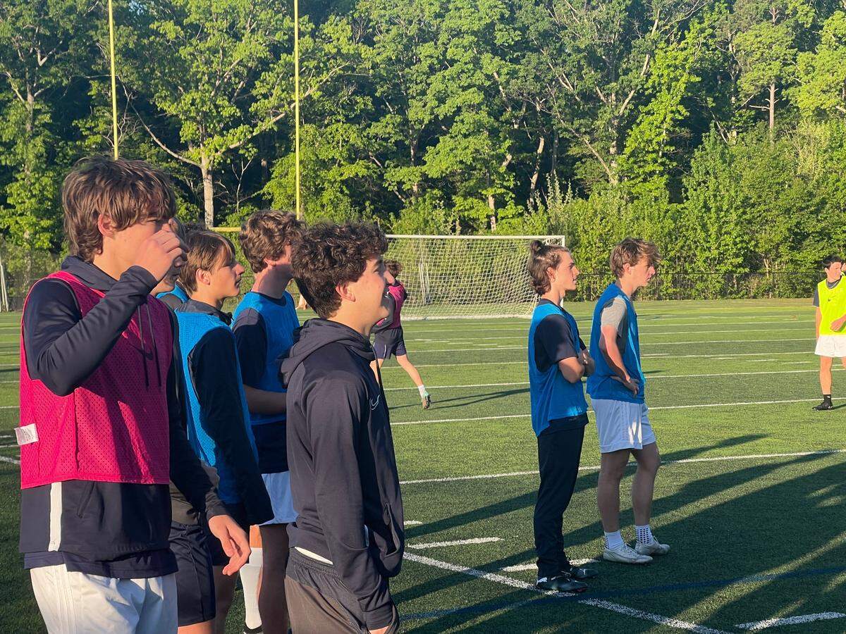 Clover defender Blake Harrill (second blue training jersey from right, arms folded), midfielder Cory Lownds (middle, black hoodie), and forward James Jenkin (left, pink training hoodie) watches Clover players train from the sidelines at a morning practice.